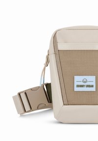 Bézs és drapp keresztpántos táska texturált anyagból, állítható pánttal és logófolttal. Masszív, téglalap alakú kialakítás jellemzi.