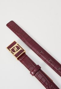 Bordeaux lærrem med krokodillemønster, gullfarget spenne med logo, og holder med hull for justering.