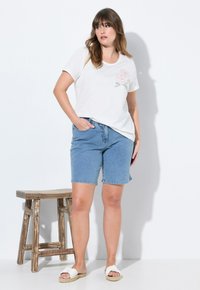 Camiseta blanca con bordado floral rosa, combinada con pantalones cortos de mezclilla azules. La modelo lleva sandalias blancas con tiras. Diseño sencillo sobre un fondo liso.