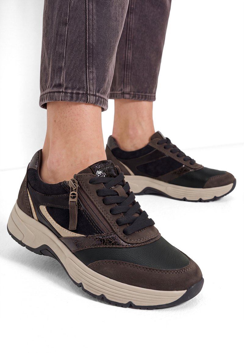 Άτομο που φοράει καφέ και μαύρα δερμάτινα sneakers με κορδόνια και φερμουάρ, συνδυασμένα με γυρισμένο σκουρόχρωμο παντελόνι, στέκεται σε λευκή επιφάνεια.