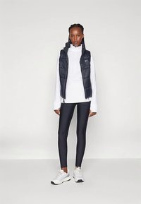 Chaleco acolchado en azul marino oscuro sobre una camiseta blanca de manga larga, combinado con leggings negros y zapatillas blancas, con cierre de cremallera y bolsillos.