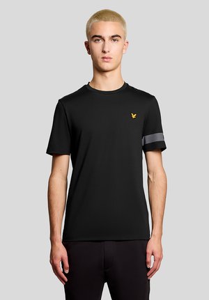 Ung mand med kort blondt hår iført en sort T-shirt med en gul fuglelogo på brystet og en grå stribe på venstre ærme.