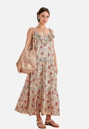 Mujer con un vestido largo sin mangas de flores con volantes, llevando un gran bolso tote tejido y usando sandalias y joyería turquesa.