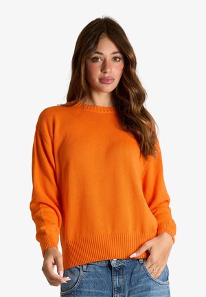 Donna con lunghi capelli mossi castani che indossa un maglione arancione di maglia e jeans blu, in piedi con una mano in tasca su sfondo bianco.