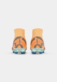 Des chaussures de football orange vif présentent un design montagnard avec des motifs texturés et une semelle extérieure bleue transparente. Marque visible sur le talon.