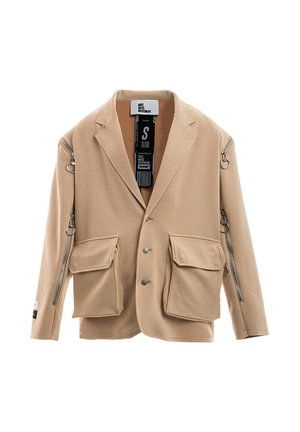 Joven con rastas que lleva una chaqueta beige de gran tamaño con cremalleras, pantalones sueltos a juego, camisa de malla y botas blancas con cordones, de pie sobre fondo blanco.