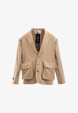 Joven con rastas que lleva una chaqueta beige de gran tamaño con cremalleras, pantalones sueltos a juego, camisa de malla y botas blancas con cordones, de pie sobre fondo blanco.