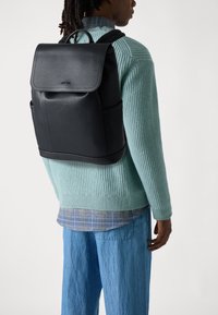 Black leather backpack with a flap closure, two side pockets, and padded shoulder straps. Smooth texture and minimalistic design.