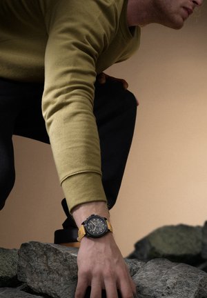 Montre chronographe noire avec un bracelet jaune, dotée de cadrans circulaires et d'une surface texturée. Le porteur se trouve sur une surface rocheuse.
