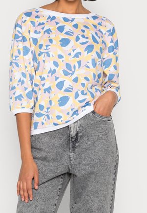 Mujer con camiseta de manga larga de estampado pastel con formas florales abstractas en azul, amarillo y blanco combinada con vaqueros grises lavados.