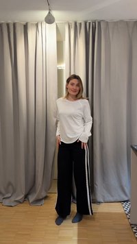 Jeune femme en chemise blanche à manches longues et pantalon noir avec des bandes blanches, debout pieds nus sur un sol en bois devant des rideaux beige.