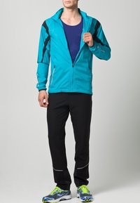 Leichte türkisfarbene Sportjacke mit schwarzen Akzenten und Reißverschluss, kombiniert mit schwarzen Hosen und Laufschuhen, die blaue und grüne Details aufweisen.