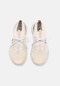 Beige nät träningsskor med vita snören och spräckliga grå sulor. Har en texturerad överdel och logotyp på tungan och hälen.