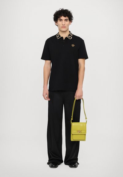 Polo shirt nera con un design del colletto geometrico a contrasto, abbinata a pantaloni larghi neri e una piccola borsa gialla con un accento dorato.