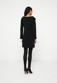 Sieviete stāv pret baltu fonu, tērpusies melnā garroku kleitā, melnās zeķbiksēs un melnās augstpapēžu kurpēs, redzama no muguras.