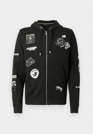 Melnas krāsas kapucis ar rāvējslēdzēju, izgatavots no mīksta auduma, rotāts ar dažādām balti izšūtām uzlīmēm. Ir pievienotas aukliņas, kā arī ribotie aproces un apakšmala.