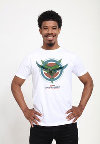 Camiseta blanca con un gráfico colorido de un personaje volando con alas, rodeado de diseños circulares y triangulares en verde y marrón.