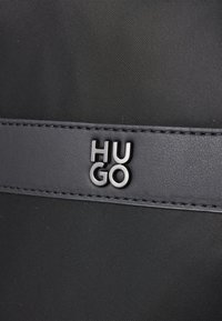 Sac en tissu noir avec une bande en cuir lisse arborant le logo embossé "Hugo" en métal argenté. Surface texturée, design élégant.