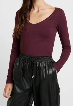 Femme portant un haut manches longues col en V bordeaux profond et un pantalon noir, brillant, taille élastique avec cordon, une main dans la poche.