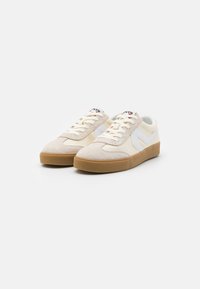 Zapatillas de ante marrón claro y cuero blanco roto. Presentan una suela de goma, parte superior texturizada y un logo lateral en contraste.