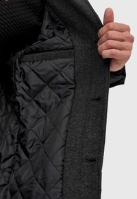 Innenseite eines grauen Hahnentritt-Mantels mit einer schwarz gesteppten, diamantförmigen Innenfütterung, die eine Brusttasche und strukturierte Stoffe zeigt. Hand, die den Mantel öffnet.