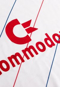 Witte stof met vette rode tekst die "Commodore" leest, met een gestileerd rood logo dat lijkt op een "C" en twee driehoekige accenten. Blauwe lijnen kruisen elkaar.