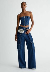 Denim kombinacija koja uključuje bezrukavnu bluzu sa zip zatvaračem i široke cargo hlače s više džepova. Srebrna crossover torba s detaljima od lanca.