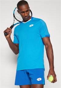 Blue athletic t-shirt with short sleeves and a white logo. Worn with matching blue shorts. Holding a tennis racket and ball.