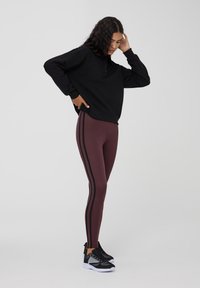 Felpa nera con mezza zip e bottoni, abbinata a leggings bordeaux con strisce laterali nere e scarpe da ginnastica nere.