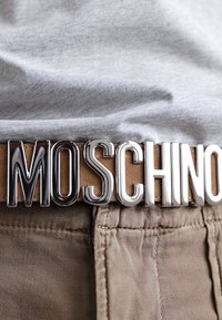 Barna bőrből készült öv nagy, fényes ezüst fém csattal, amelyen a "MOSCHINO" felirat olvasható, szürke ing és khaki nadrág előtt bemutatva.