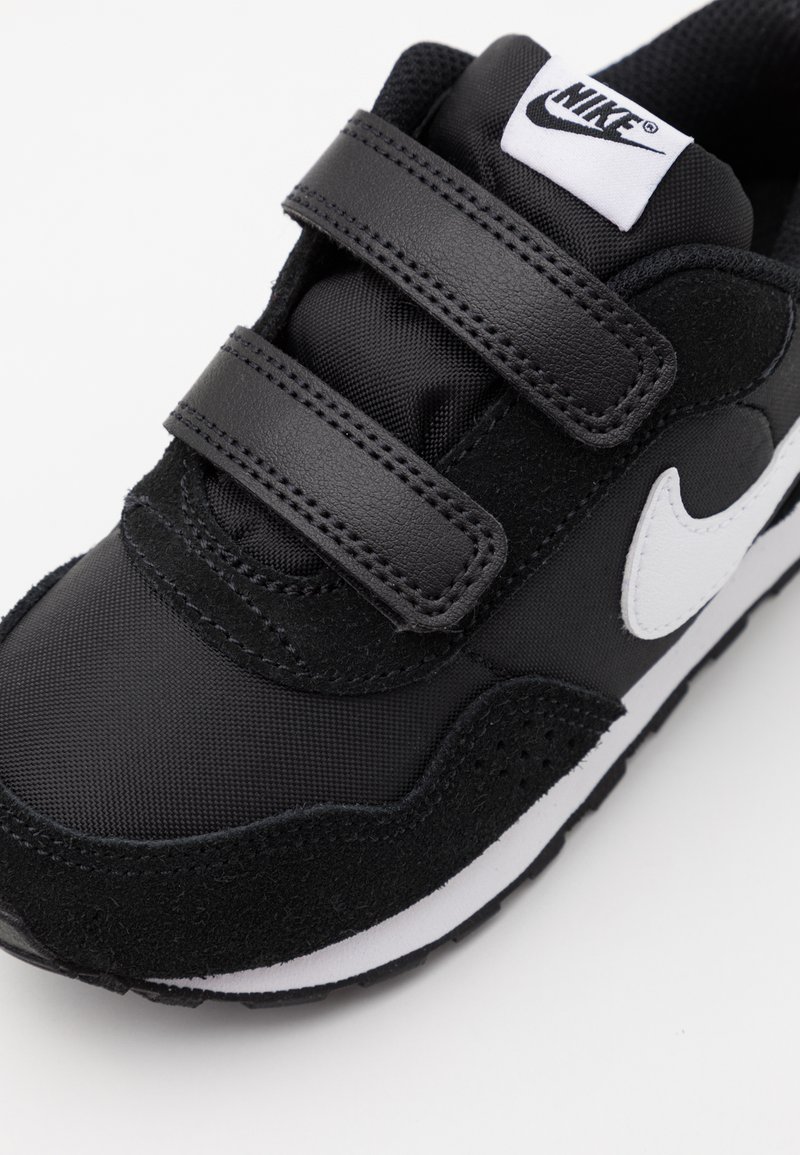 Zapatilla deportiva negra que presenta materiales de ante y sintéticos, correas de Velcro, logo de swoosh blanco y una entresuela blanca sobre una suela texturizada.