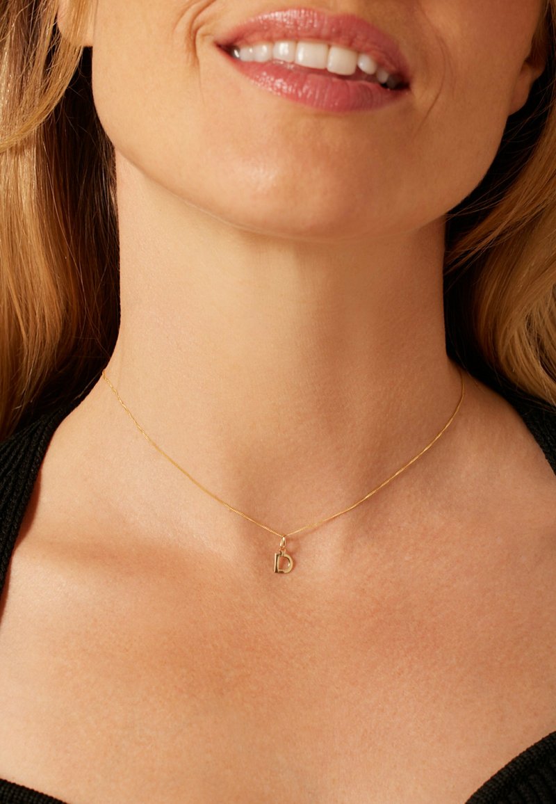 Gold necklace featuring a delicate chain and a small, textured letter "D" pendant, set against bare skin.