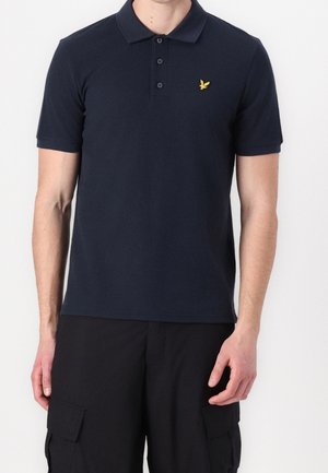 Mężczyzna w granatowej koszulce polo z krótkim rękawem, z trzema guzikami i małym żółtym logo ptaka na piersi, ubrany w czarne szorty bojówki.