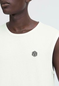 Maglietta senza maniche grigia chiara con scollatura rotonda, caratterizzata da un piccolo logo geometrico ricamato nero sul petto; tessuto morbido e liscio.
