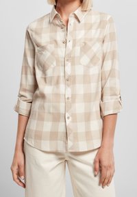 Femme portant une chemise à manches longues à carreaux beige et blanche avec les poignets retroussés et un pantalon couleur crème, vue du cou aux hanches.