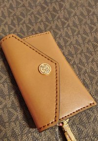 Portafoglio in pelle marrone con finitura texturizzata, caratterizzato da un emblema dorato e cuciture a contrasto lungo i bordi. Design compatto e angolato.
