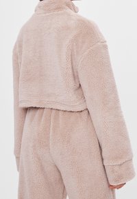 Krótka, miękka kurtka z polaru w jasno beżowym kolorze z wysokim kołnierzem, szerokimi rękawami i puszystą fakturą. Charakterystyczne przeszycia na ramionach.