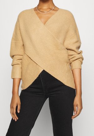 Femme portant un pull cache-cœur en maille côtelée beige avec manches longues et un pantalon noir taille haute, assorti d’un collier chaîne dorée.