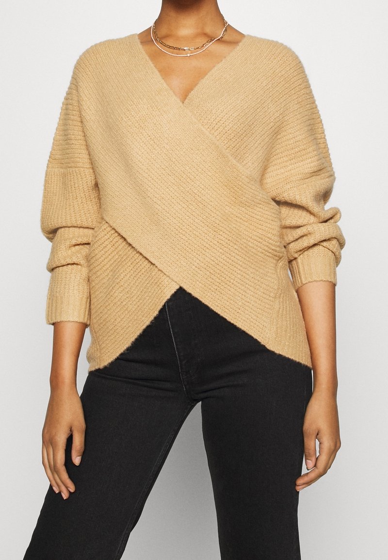 Femme portant un pull cache-cœur en maille côtelée beige avec manches longues et un pantalon noir taille haute, assorti d’un collier chaîne dorée.
