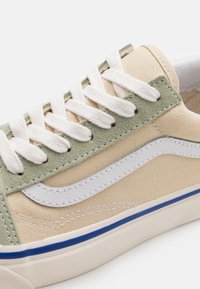 Baskets en toile beige avec lacets blancs, empiècement à œillets en daim vert, bande latérale blanche et semelle crème bordée de bleu.
