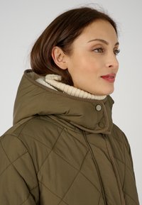 Chaqueta acolchada color caqui con un alto cuello y una capucha sobredimensionada, con un suave forro interior beige y cierre de botones a presión.