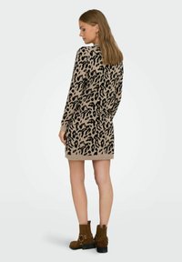 Femme de dos portant une robe mini à manches longues en imprimé léopard beige et noir avec des bottes à la cheville marron sur un fond blanc uni.