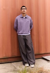 Lys lilla genser med glidelås og krage, kombinert med løse grå bukser og svart-hvite joggesko. Står mot en rustfarget vegg.