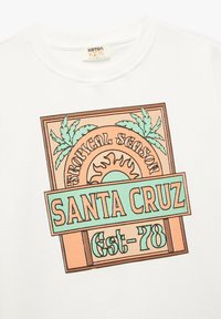 Camiseta blanca de algodón con un tejido texturizado. Presenta un gráfico colorido con "SANTA CRUZ" y hojas de palmera tropicales.
