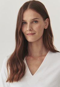 Top bianco a V con vestibilità rilassata. Il modello ha lunghi capelli castani ondulati e occhi blu. Tessuto morbido, design minimalista, sfondo neutro.