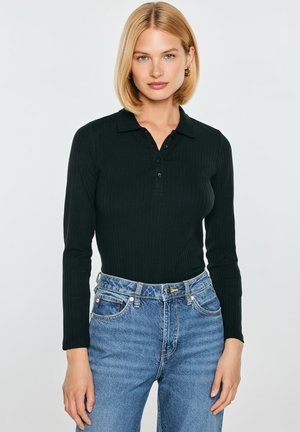 Kobieta o blond bob fryzurze, ubrana w czarną prążkowaną koszulkę polo z długim rękawem i niebieskie jeansy z wysokim stanem, stojąca na białym tle.