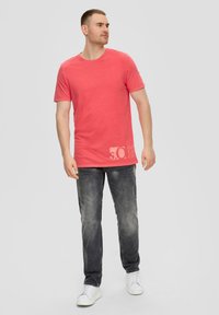Korallefarbenes T-Shirt mit kurzen Ärmeln und Rundhalsausschnitt, ausgestattet mit einem verblassten Grafikdruck und kombiniert mit hellgrauen Jeans und weißen Sneakers.