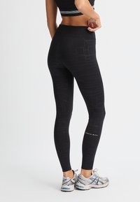 Leggings neri con motivo geometrico texturizzato, vita alta e dettaglio con logo. Abbinati a scarpe da ginnastica grigie con accenti blu.