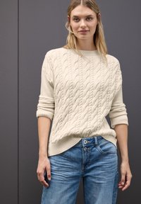 Cremefarbener Kabelstrickpullover mit geripptem Saum und Bündchen, kombiniert mit hellblauen Jeans aus Denim mit doppeltem Knopfverschluss und sanften Verfärbungen.