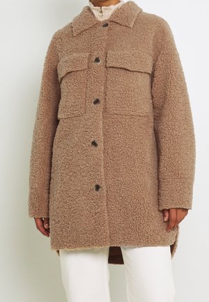 Brązowy, teksturowany płaszcz teddy zapinany na guziki z dużymi kieszeniami na piersiach, noszony na białych spodniach i beżowym golfie.